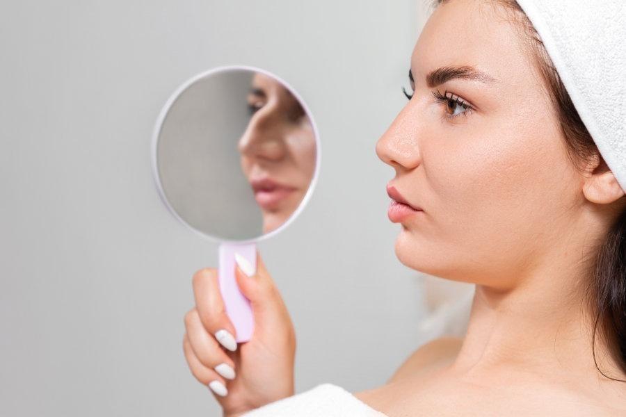Rhinoplasty. A young beautiful woman holds a mirror with aquiline nose. Comparison of results before and after plastic surgery.