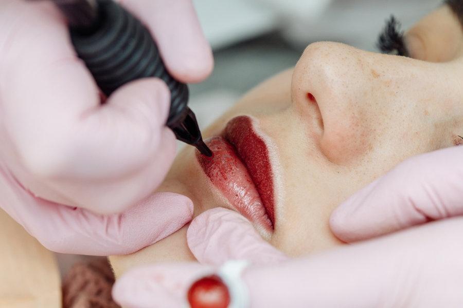 Woman having permanent makeup on lips in beauty salon. Cosmetologist in gloves applying tattoo with machine, macro view